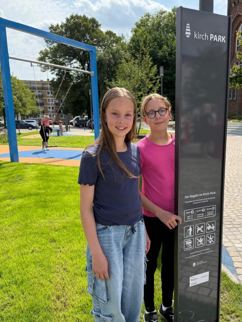 Zwei Mädchen neben dem Schild "kirch Park" vor einer Schaukel.